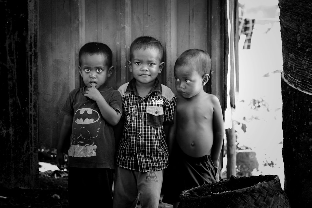 A black and white photo of three Malay children standing next to each other, one holding his chin with his hand while the two on either side smile at the camera, wearing a Batman t-shirt and long pants outside their house in a kampung setting, taken in the style of Hasselblad H2D. –ar 128:85