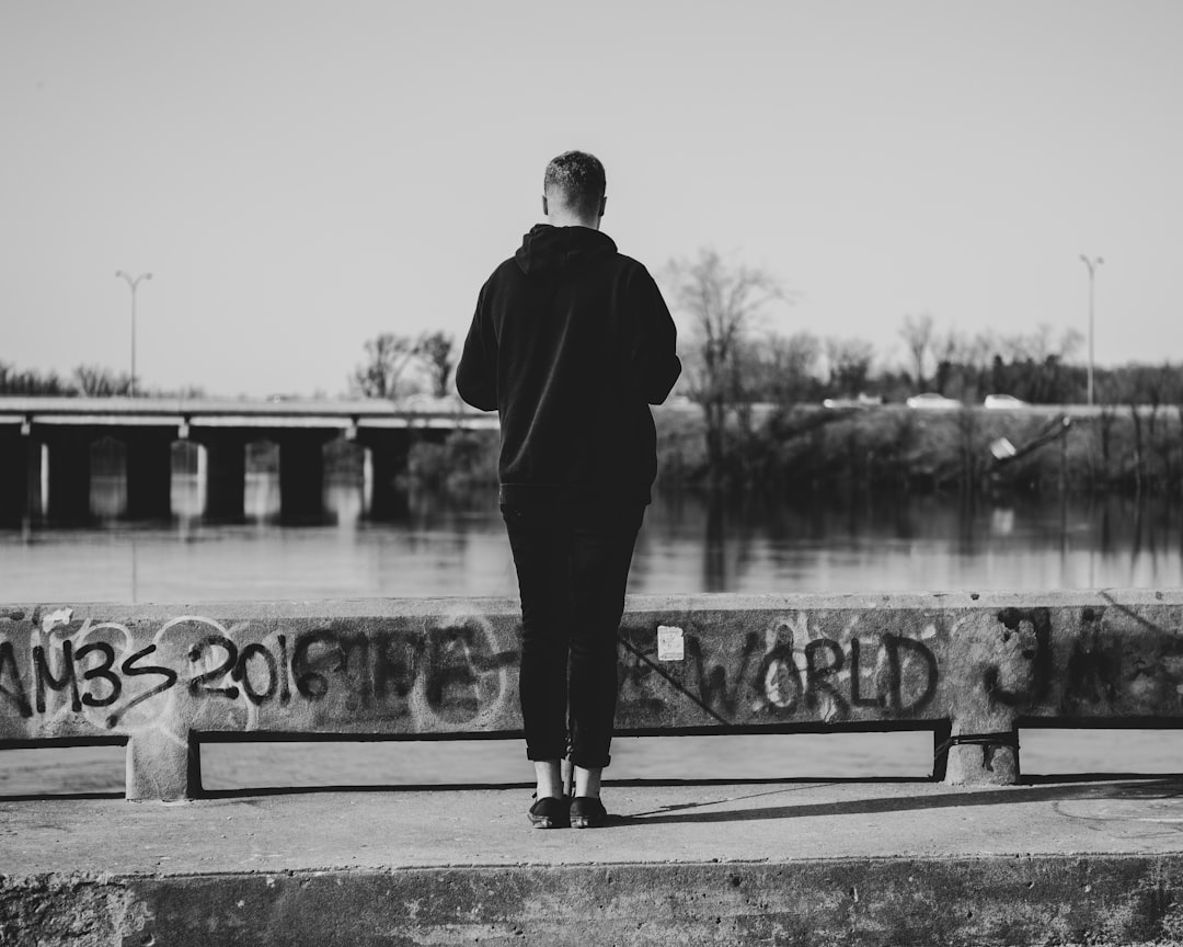 A black and white photo of an adult male standing on the edge of concrete, wearing a dark hoodie with his back to the camera, looking at water with graffiti “2036” in front, a bridge over a river behind him, taken with a Sony Alpha A7 III in the style of street photography. –ar 64:51