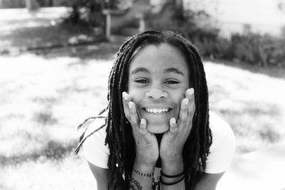 black and white portrait of happy black teenage girl with dreadlocks, hands on face, posing for senior pictures in the backyard, shot from above --ar 128:85