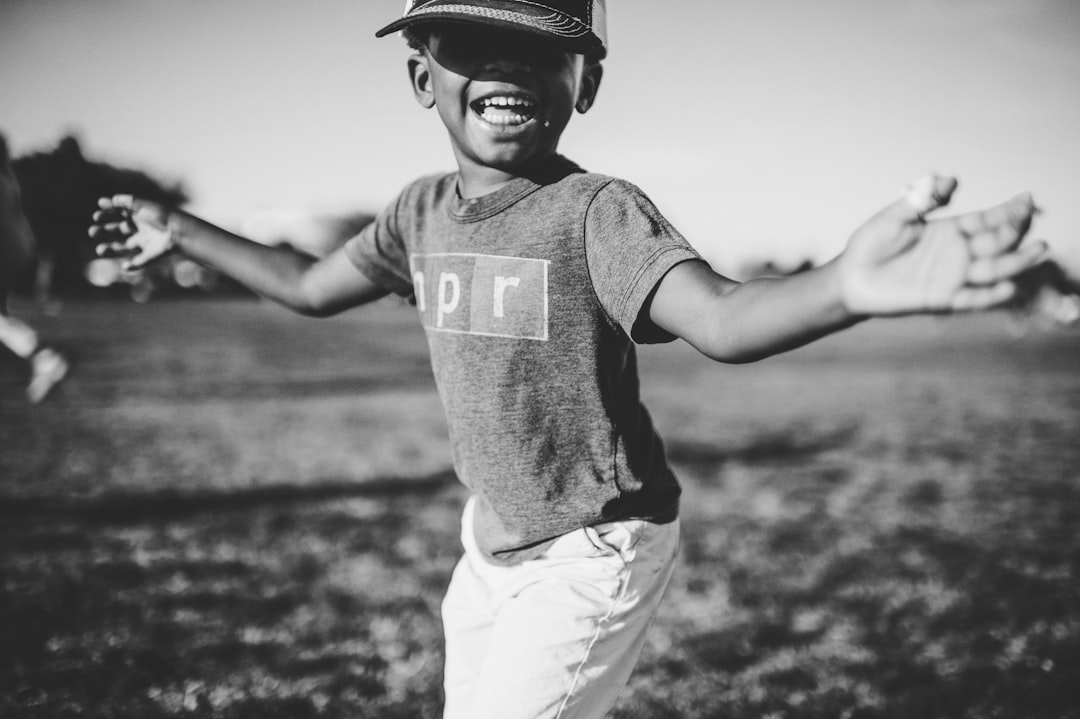 Black and white photo of a happy smiling boy wearing a baseball cap and T-shirt with “Bold PDave” written on it, hands outstretched in the air, dancing in an open field with a blurred background and sunlight casting soft shadows. Taken with a Nikon Z7 II camera, 50mm lens at f/2.8 aperture. Black children playing sports outdoors. Use soft focus to emphasize facial expressions and emphasize his joyful expression through natural light. The scene captures him having fun while celebrating victory in the style of soft focus photography. –ar 128:85