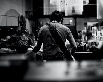 Photo of the back view, from behind, of an American man in his thirties with dark hair and wearing jeans standing at a restaurant counter playing guitar for people eating inside the restaurant. Black and white, in the style of Hasselblad photography, shallow depth of field, highly detailed photo, hyper realistic. --ar 64:51