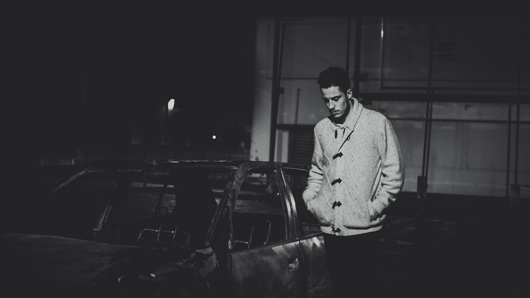 Black and white street photography of a young man wearing a wool coat standing next to a car in a dark warehouse at night time with low light in the grainy film noir style. Grainy black and white photography with a 35mm lens at f/2.8 with accent lighting and global illumination, shot on Kodak Gold 400 film in the style of film noir. –ar 16:9