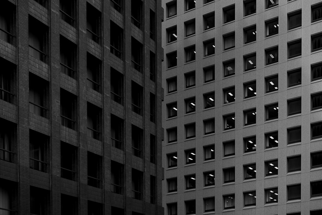 A monochromatic photograph of an office building with many windows, each window glowing from the inside and creating intricate patterns on its exterior walls. The photo captures the interplay between light and shadow in the urban setting, emphasizing architectural details and textures. Black background. The photograph was taken with a Sony Alpha A7 III camera using a Zeiss Batis lens at an aperture setting of f/2.8 in the style of minimalist architectural photography. –ar 128:85