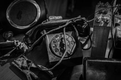 Black and white photograph of an old, dusty telephone with its wires tangled up on the table in front of it, surrounded by other vintage items from early photography equipment to classic musical instruments. The focus is sharp on the phone's number line, capturing intricate details that highlight textures like leather or metal. Soft lighting creates shadows around the scene, adding depth and character to each object. --ar 32:21