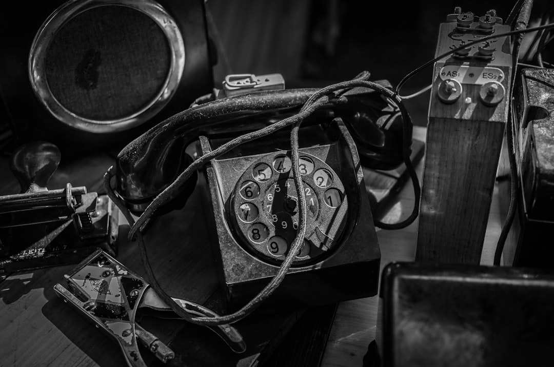 Black and white photograph of an old, dusty telephone with its wires tangled up on the table in front of it, surrounded by other vintage items from early photography equipment to classic musical instruments. The focus is sharp on the phone’s number line, capturing intricate details that highlight textures like leather or metal. Soft lighting creates shadows around the scene, adding depth and character to each object. –ar 32:21