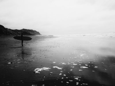 Black and white photography of a surfer standing on the beach holding his surfboard. The beach has black sand. The marine life at Kanap Schanck Beach is lush, in the style of minimalism. --ar 4:3