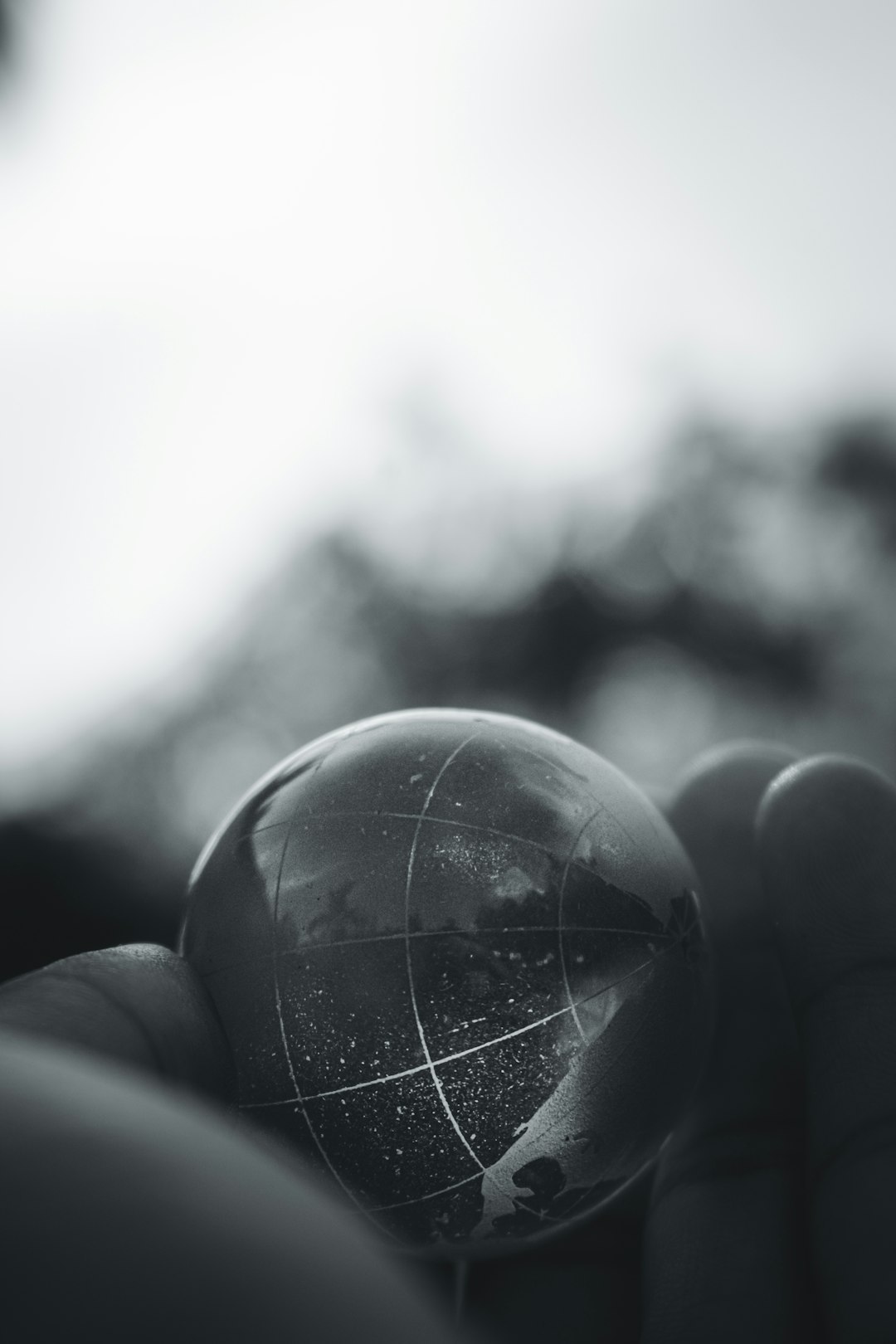 A closeup of a glass globe held in the hand, with mountains and rivers visible inside. The background is blurred to emphasize the foreground object. The photography style is black and white. High contrast between light and dark tones creates strong texture. Soft side lighting highlights details on the earth’s surface. Shot from an overhead perspective using a Nikon D850 camera. –ar 85:128