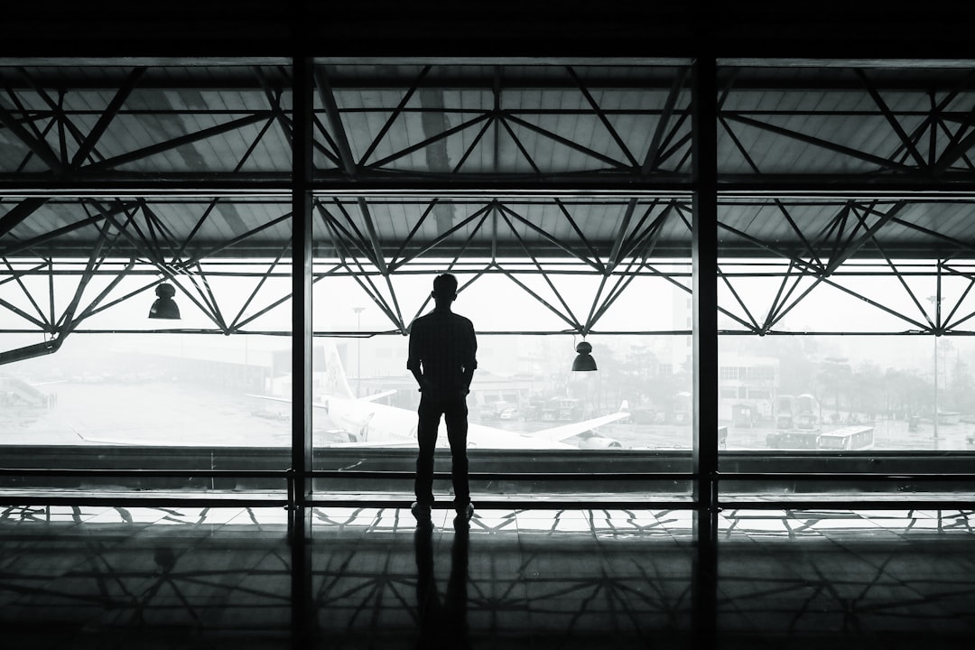 Silhouette of man standing in airport terminal, airplane visible through window, alone and contemplative mood, monochrome, minimalist style, high resolution, –ar 128:85