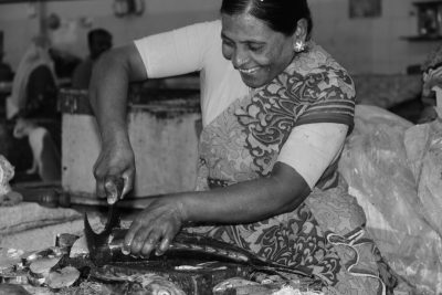 A black and white photograph shows an Indian woman in her late thirties, wearing traditional attire, smiling as she carves fish at the market's counter top. She has dark hair with a high ponytail tied back into two braids that flow down to either side of her head. The scene captures her skillful handiwork against the backdrop of various tropical fish being sliced open on large wooden counter tops covered in plastic wrap. The photograph is in the style of a traditional Indian photographer. --ar 128:85