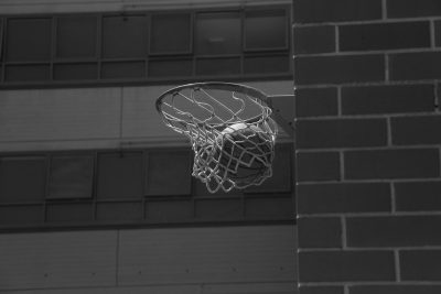 A basketball rim and net on the wall of an indoor gym, captured in black and white photographic style. The scene is framed from below, focusing only on the hoop with no ball visible. Soft lighting casts gentle shadows across the brick surface, highlighting details like the wire mesh around the basket and subtle textures on its surroundings. This composition creates a sense of classic sports action against a solid background, emphasizing contrast between lightness and darkness. --ar 128:85