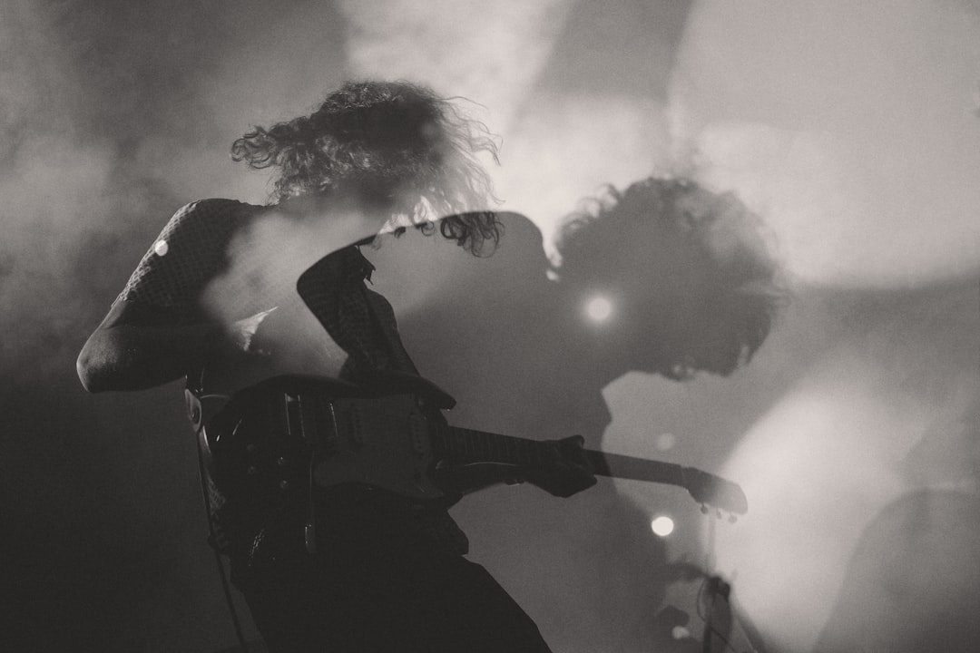 Black and white photography shows two young men playing guitar on stage. The male lead wears an open black shirt with curly hair blowing in his face while performing for thousands at a makeup themed concert. The other man plays electric bass and wears long sleeves. The scene takes place from behind them on stage. Smoke fills around their heads. They have very messy hairstyles. Stage lighting creates a soft glow around both faces, creating a moody atmosphere. –ar 128:85