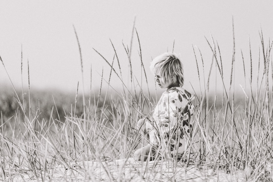 Black and white photograph of an adorable little boy with short blond hair, wearing pajamas covered in floral patterns, sitting on the beach dunes surrounded by tall sea grasses. The scene is captured from behind as he gazes out at the ocean. Soft natural lighting enhances his features. –ar 128:85