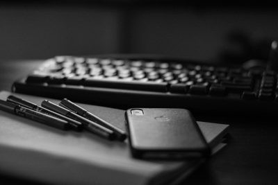 Black and white photograph of an iPhone, pen on the desk next to it, keyboard in the background, high contrast, low light, cinematic, shot in the style of Sony Alpha A7 III --ar 128:85