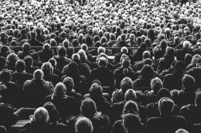 A large crowd of people in the stands at an outdoor stadium, watching and listening to someone giving their view on something during an event. Shot in the style of Nikon D850 with an 24mm f/3.6 lens. Background is black, lighting is natural, skin is hyper realistic, colors are muted, film grain effect is applied, low contrast black & white filter is used. --ar 128:85