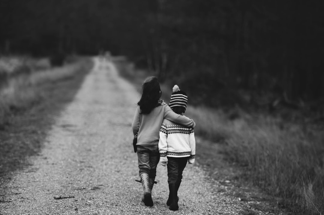 Black and white photo of two young children walking down an empty country road. One child is holding the other’s hand as they walk towards the camera. The first has long dark hair wearing jeans and a sweater with tall boots on her feet. She wears black leggings underneath her trousers. Both wear striped hats. Shot from behind them in a wide angle view, capturing their full bodies and the open space around them. The scene conveys freedom and connection between family members in the style of [Ansel Adams](https://goo.gl/search?artist%20Ansel%20Adams). –ar 128:85