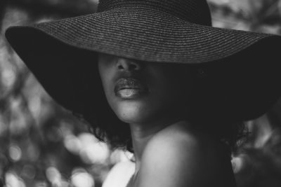 Black and white portrait of an African American woman wearing a large hat, with a focus on her lips. She is outdoors in nature, surrounded by trees. The lighting creates soft shadows that accentuate the contours of her face. Shot with a Leica M6 using Ilford HP5 film, in the style of the photographer. --ar 128:85