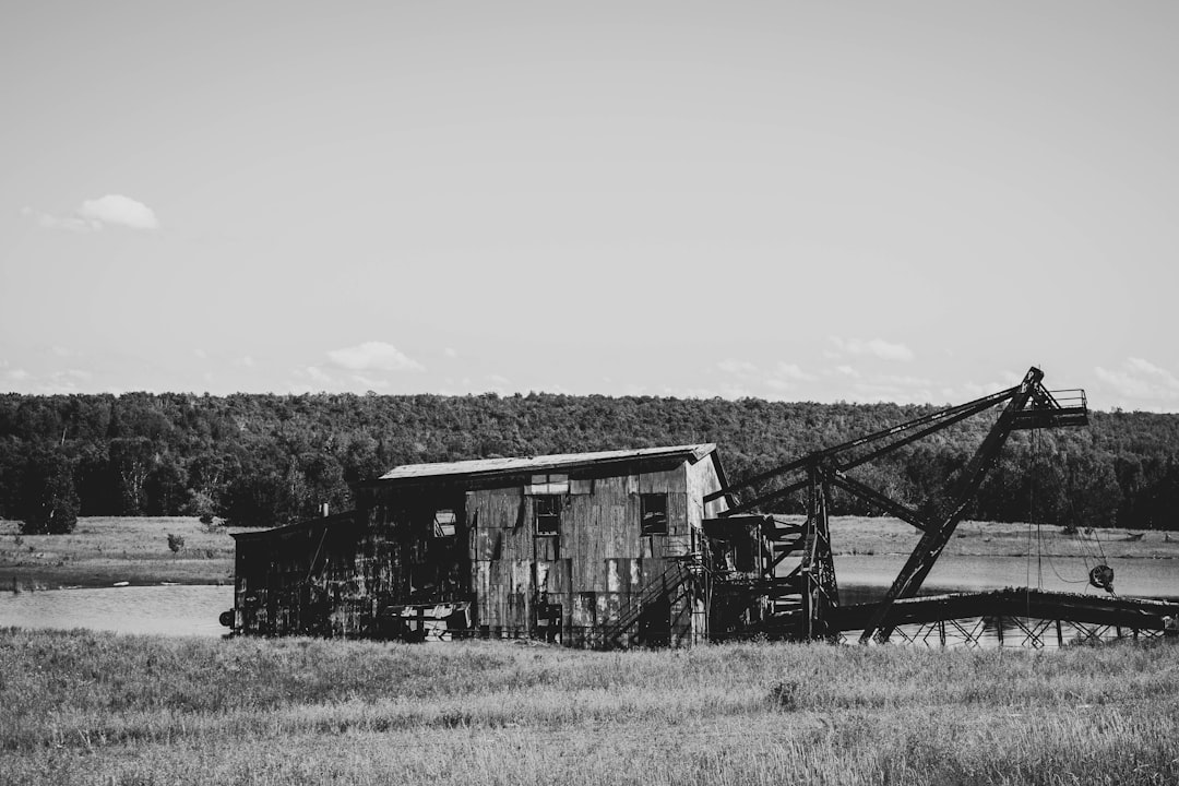 black and white photo of an old mining machine in the middle distance, next to it is a small wooden building on wheels with cranes coming out from its roof, in front there’s grassy field, hills covered by trees far away, sunny day, natural light, raw style –ar 128:85