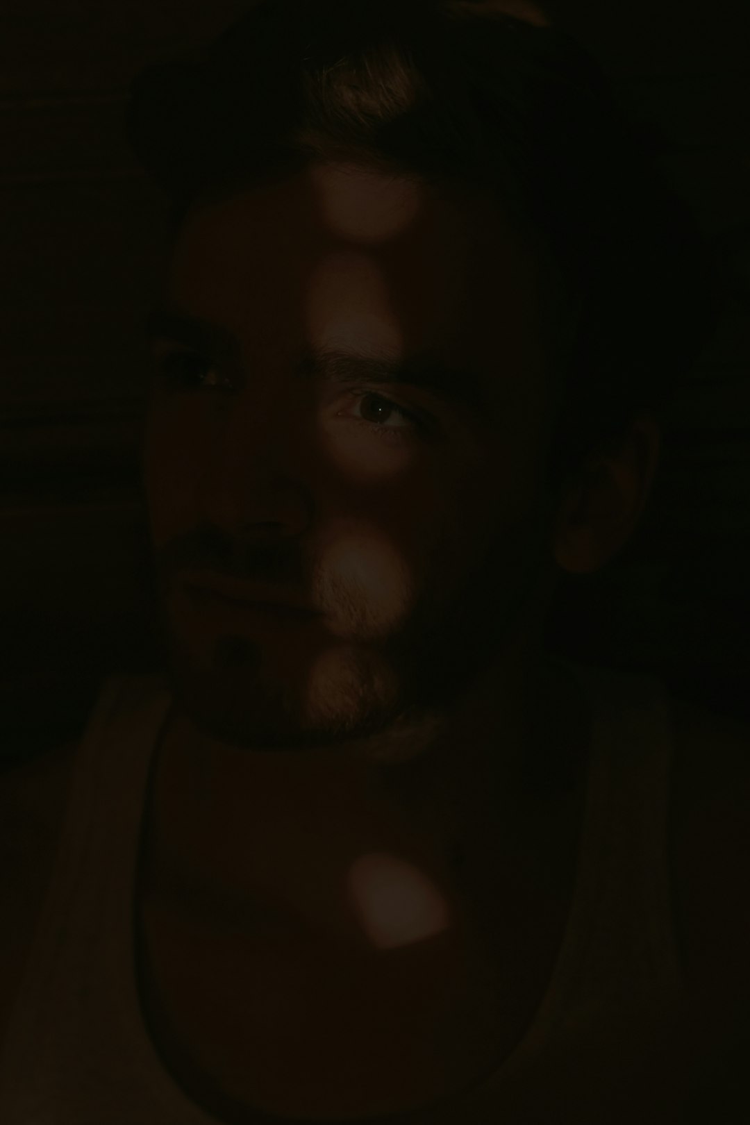 A dark portrait photograph of an attractive man, illuminated by the soft glow from his phone screen. The lighting creates dramatic shadows and highlights in his face, giving it depth and dimension, highlighting facial features like eyes, nose, lips and skin texture. His hair is black with some brown on one side. He has no beard or mustache. It’s a closeup shot, focusing only on him. He wears glasses. His expression seems thoughtful as he gazes at something outside the frame, in the style of a portrait that could have been painted by a Renaissance artist. –ar 85:128