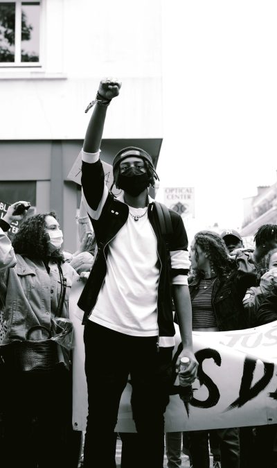 A black and white photo of an African American man in his mid-20s, wearing streetwear standing on the side with his fist raised up at a Black History Month protest surrounded by people holding signs saying "Vault Center", shot from eye level, in Paris, captured using a Canon EOS R5 mirrorless camera with a standard lens. --ar 75:128