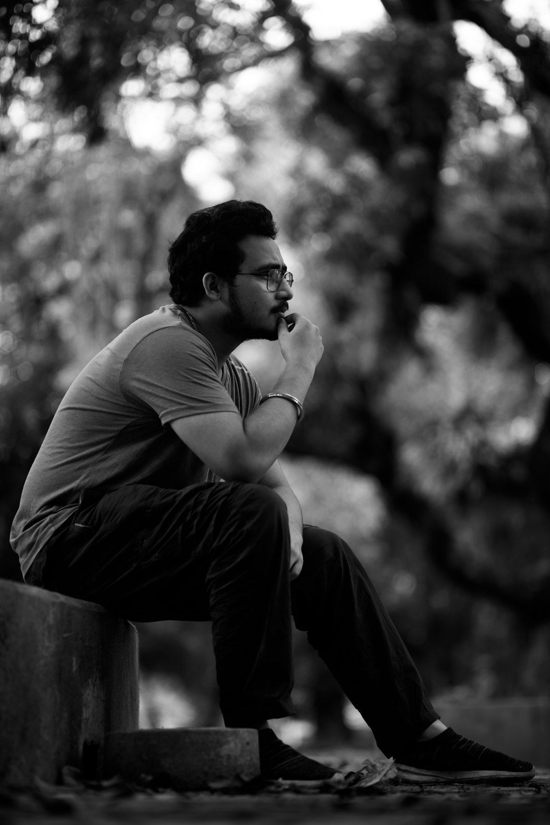 A black and white photo of an Indian man sitting on the edge of a park bench, wearing glasses with his hand on his mouth thinking about something while looking away from the camera in profile. He is sitting at a park bench, wearing a t-shirt and jeans, in a side view, full body shot with natural lighting in the style of a raw style stock photography image. –ar 85:128