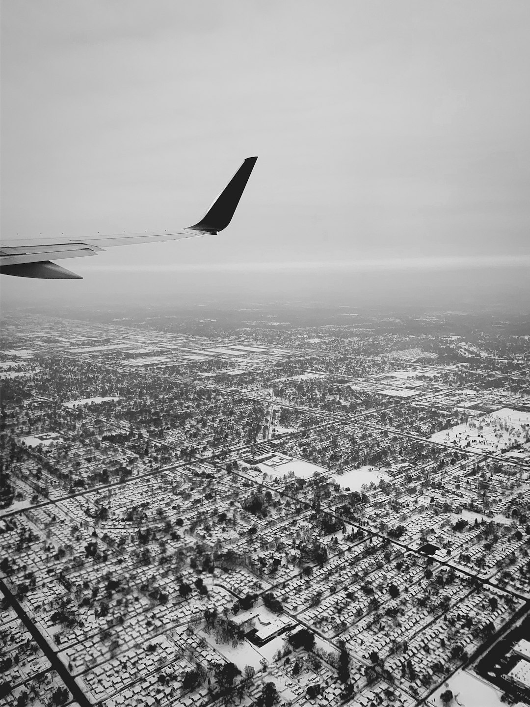 Aerial view of the snow-covered city from an airplane window, with black and white tones. The plane wing is visible in one corner, adding to the perspective. Snow covers buildings, streets, and greenery, creating a stark contrast against the grey sky. A sense of isolation surrounds you as your journey unfolds through this winter landscape. –ar 3:4