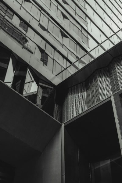 A black and white photo of the exterior architecture of an office building in London, showcasing its modern design with geometric shapes, large windows, and glass panels. The composition captures details like shadow play on walls, reflections from metal accents, and subtle textures that highlight architectural elements. Shot in the style of photographer M, with stripes, grainy texture, monochrome color scheme, striking contrast between light and dark areas, minimalistic aesthetic. --ar 85:128