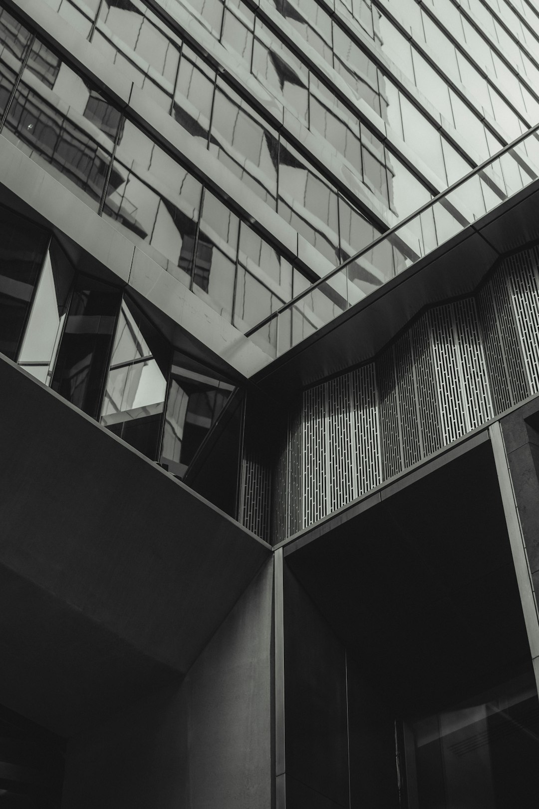 A black and white photo of the exterior architecture of an office building in London, showcasing its modern design with geometric shapes, large windows, and glass panels. The composition captures details like shadow play on walls, reflections from metal accents, and subtle textures that highlight architectural elements. Shot in the style of photographer M, with stripes, grainy texture, monochrome color scheme, striking contrast between light and dark areas, minimalistic aesthetic. –ar 85:128