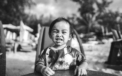 A black and white candid photo of an extremely cute happy baby sitting at the table, with messy hair, wearing a shirt with a small collar, and a dirty face from food splashes. On a beach chair in the background. The photo was taken with a Fujifilm Superia in the style of a full shot with a wide angle lens using natural light on a hot summer day. --ar 8:5