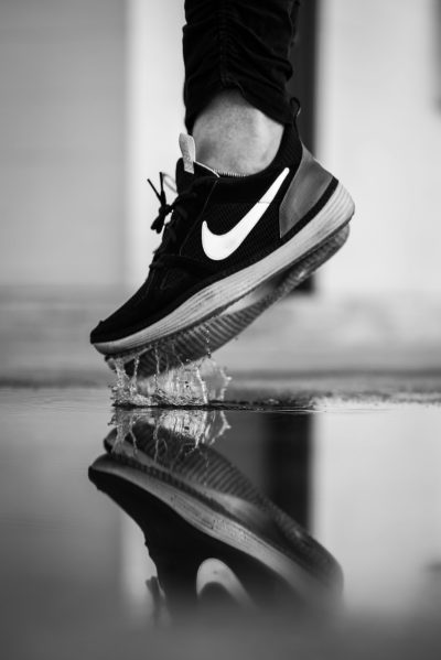 Black and white photo of the Nike running shoe on top, with water droplets in front of it. The person is wearing black pants and shoes. A reflection can be seen below. In the background, there is an empty street or floor. Captured in the style of Nikon D850 camera using a high-resolution DSLR for detailed photography. --ar 85:128