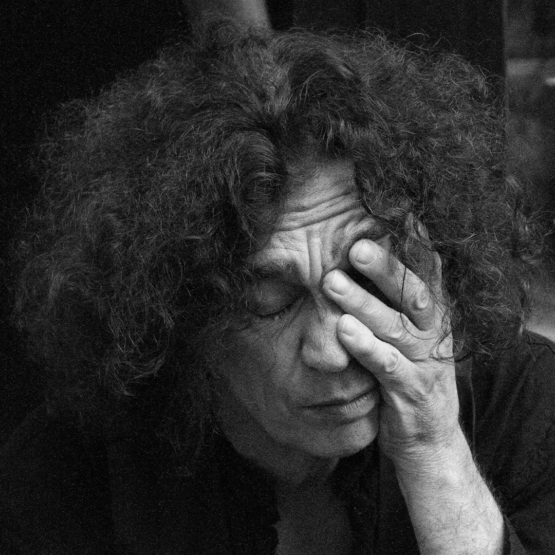 Black and white photo of Stiv Bator from The Dead Boys, with long curly hair covering his face, holding one hand to the side of his head in pain, crying in sadness, looking down at an angle in low light in a closeup.