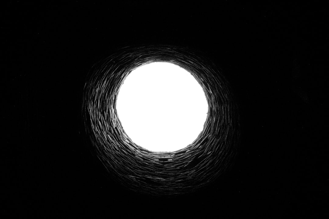 Black and white sketch of the inside view of an oval hole in darkness with light at its center, symbolizing hope or mystery. The black background adds depth to the minimalist drawing. White circle against dark interior. Minimalist art style. –ar 128:85