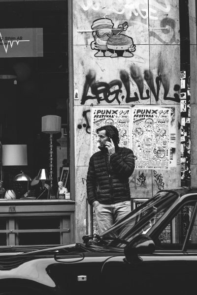 A black and white photo shows the front wall of an urban coffee shop, with graffiti saying "Iwaipunk festival". A man in his late thirties stands next to the wall talking on his phone, wearing a puffer jacket and jeans. He is leaning against one side of a car parked outside the window. The building has a modern architectural design. --ar 85:128