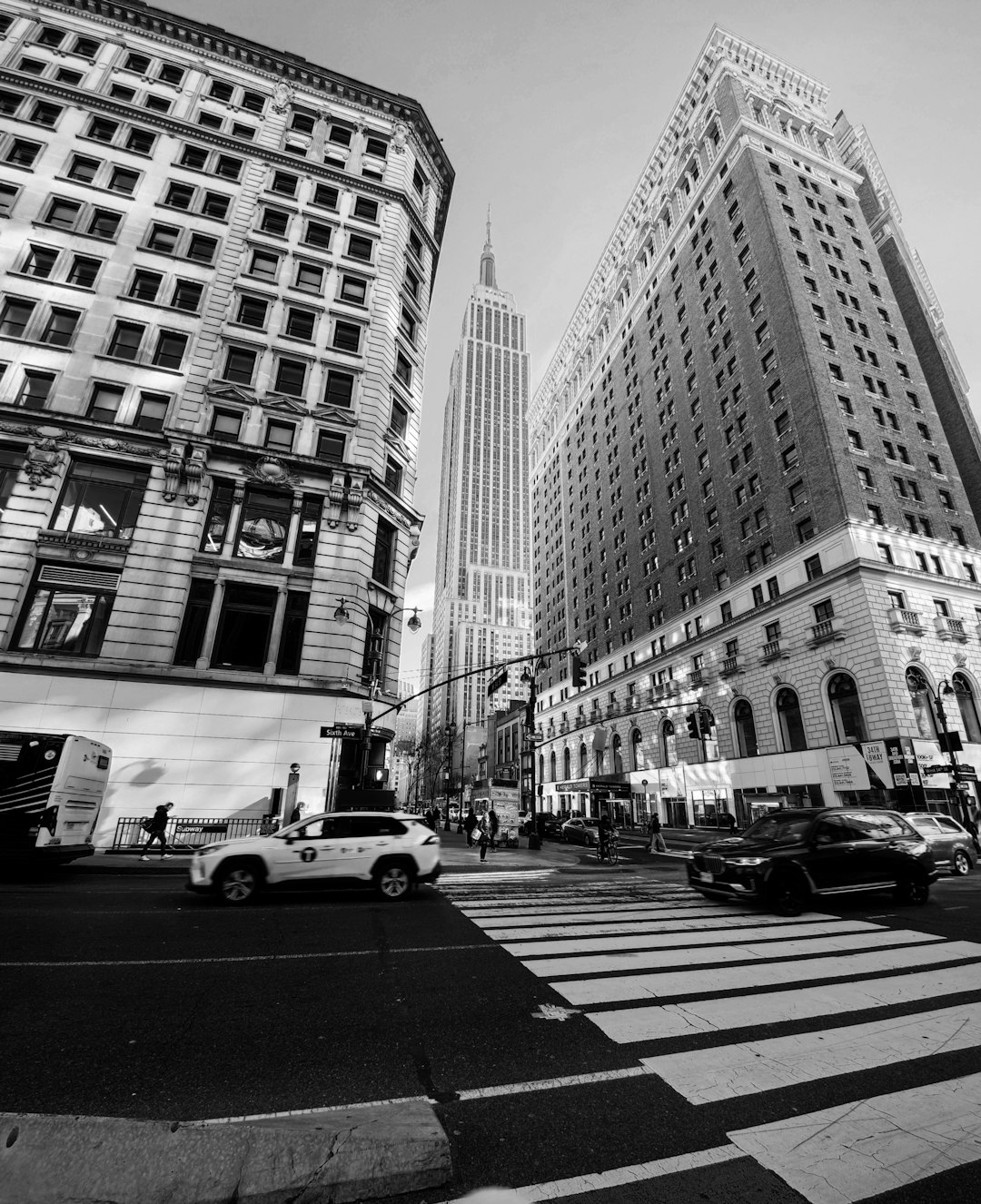 A black and white photo of the Empire State Building. A New York City street scene with buildings in the foreground and cars on the road with a pedestrian crossing in front. No people or human figures are present and there are no shadows. The photo has a low angle camera shot with a wide lens, captured in a photorealistic style in the style of a Canon EOS R5 camera. –ar 13:16