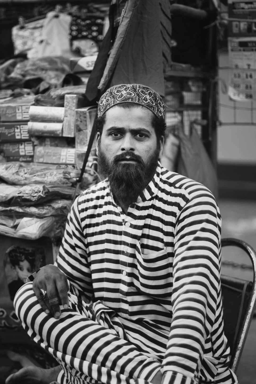 Black and white street portrait of a Bangladeshi man wearing a striped long shirt, sitting in front of a market stall selling fabric, looking at the camera, with a beard, in the style of pop art, using Fujifilm Pro 400H film stock. –ar 85:128