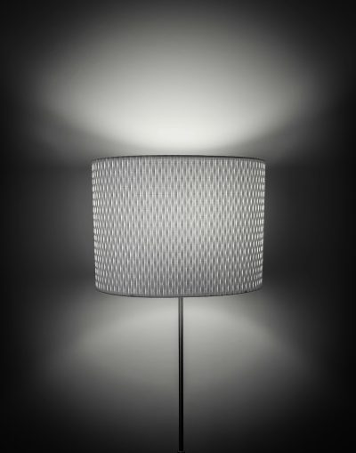 A floor lamp with a fabric shade, featuring an intricate geometric pattern in shades of grey and white. The light from the top illuminates the wall behind it, creating soft shadows that accentuate its texture. A sleek metal base adds to the modern aesthetic. High resolution photography, detailed cinematic shot, featuring cinematography in the style of modern design. --ar 25:32