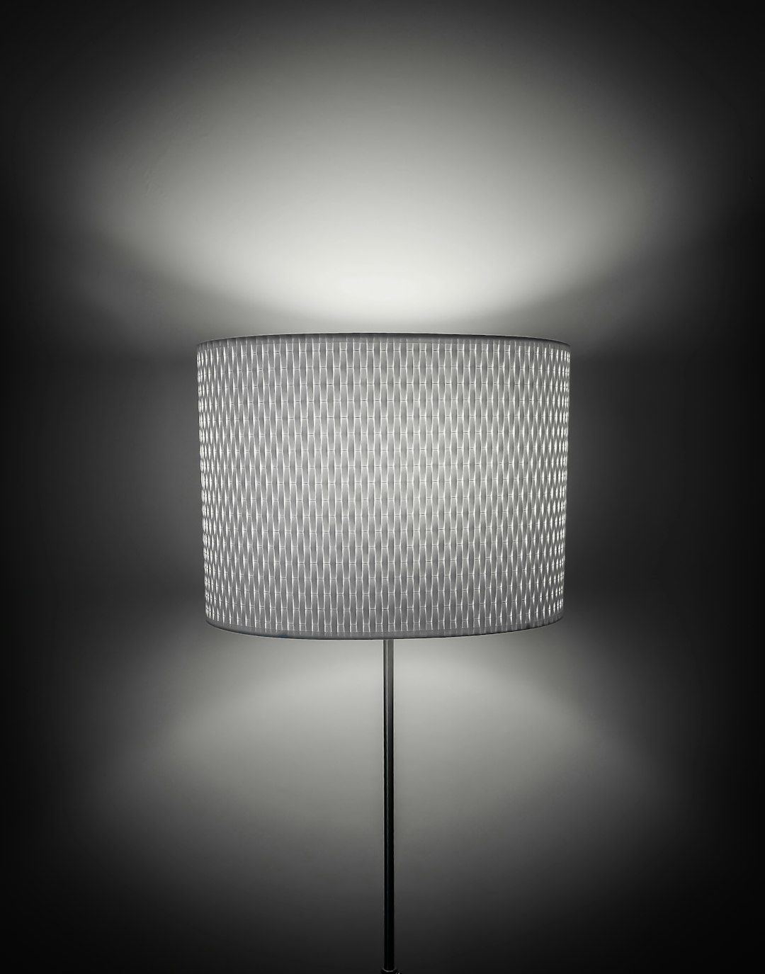 A floor lamp with a fabric shade, featuring an intricate geometric pattern in shades of grey and white. The light from the top illuminates the wall behind it, creating soft shadows that accentuate its texture. A sleek metal base adds to the modern aesthetic. High resolution photography, detailed cinematic shot, featuring cinematography in the style of modern design. –ar 25:32