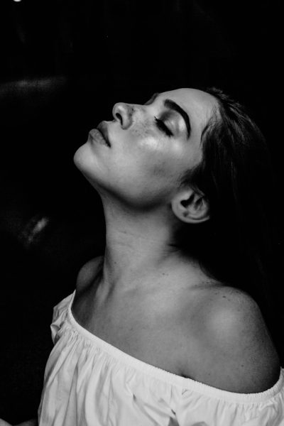 black and white photo of woman in the style of Tom OFhealth, wearing offtheshoulder shirt, her head tilted up to show skin on neck, looking upwards with eyes closed, soft lighting, low angle shot, blurred background, portrait photography, monochrome color scheme, high contrast between subject's face against dark backdrop, minimalistic composition, capturing mood and expression. --ar 85:128