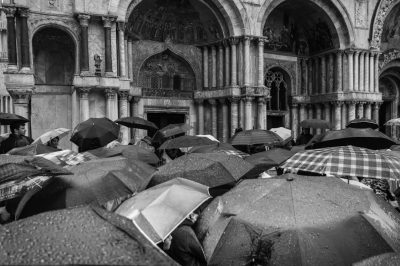 A black and white photograph of people with umbrellas in St Mark's Square, Venice during the rain on a sunny day. The scene captures an atmosphere filled with a crowd gathering under open plaid patterned umbrellas, standing around ancient architecture. The photography is in the style of high resolution photography, focusing on capturing the contrast between wet surfaces and a bright sky, with detailed attention to texture details and composition. --ar 128:85