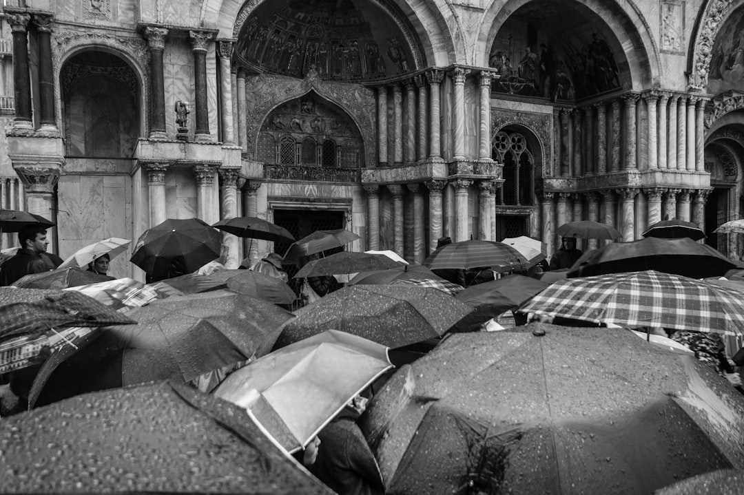 A black and white photograph of people with umbrellas in St Mark’s Square, Venice during the rain on a sunny day. The scene captures an atmosphere filled with a crowd gathering under open plaid patterned umbrellas, standing around ancient architecture. The photography is in the style of high resolution photography, focusing on capturing the contrast between wet surfaces and a bright sky, with detailed attention to texture details and composition. –ar 128:85