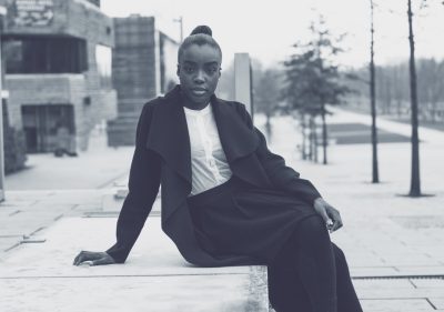 Black and white street photography of young black woman in modern outfit sitting on concrete bench, hair bun hairstyle, wearing oversized coat, posing in front of university building with contemporary architecture --ar 128:89