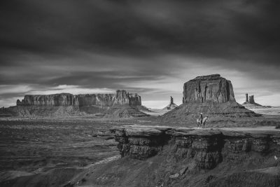 Black and white photography of a western landscape with a cowboy riding a horse in the foreground, iconic rock formations of Monument Valley, in the style of unsplash. --ar 128:85