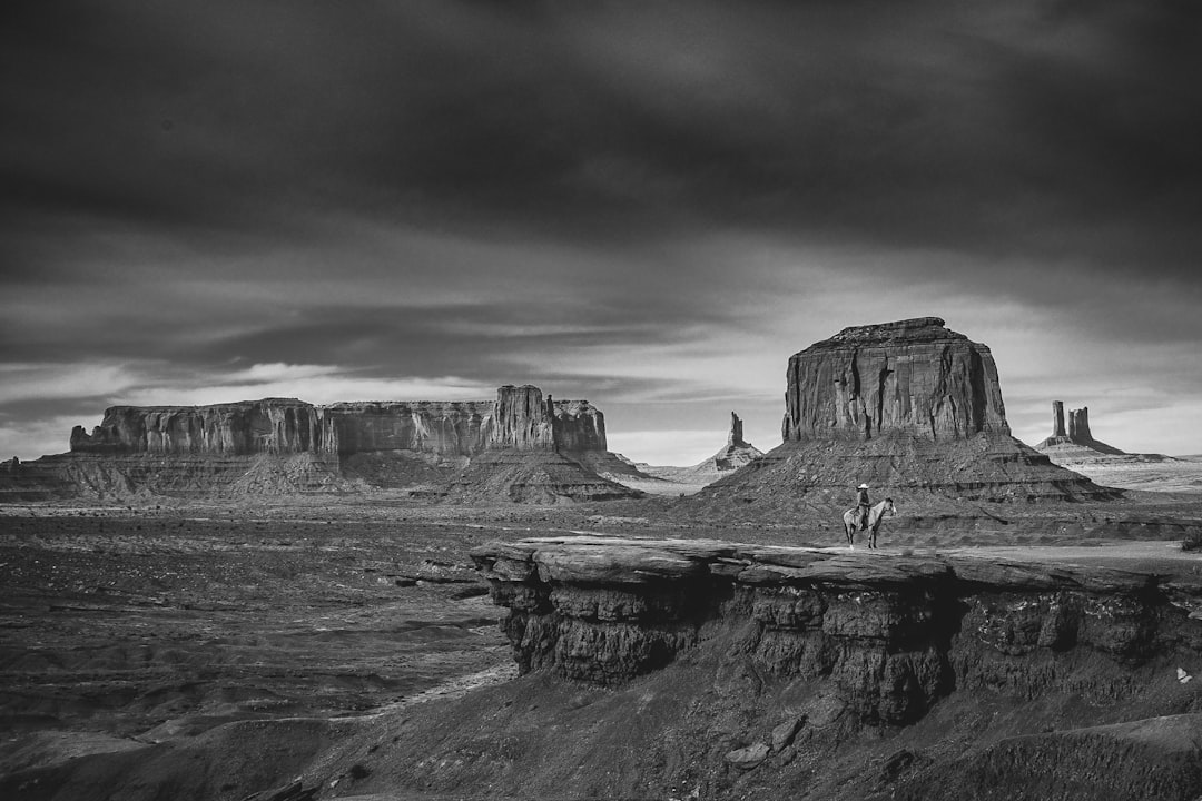 Black and white photography of a western landscape with a cowboy riding a horse in the foreground, iconic rock formations of Monument Valley, in the style of unsplash. –ar 128:85