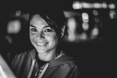 portrait of smiling woman, sitting at a bar in the evening, she is wearing a hoodie and sports , blurred background, black & white photography, shot with a Leica SL2S camera, 50mm lens, f/4 aperture, soft natural lighting. --ar 128:85