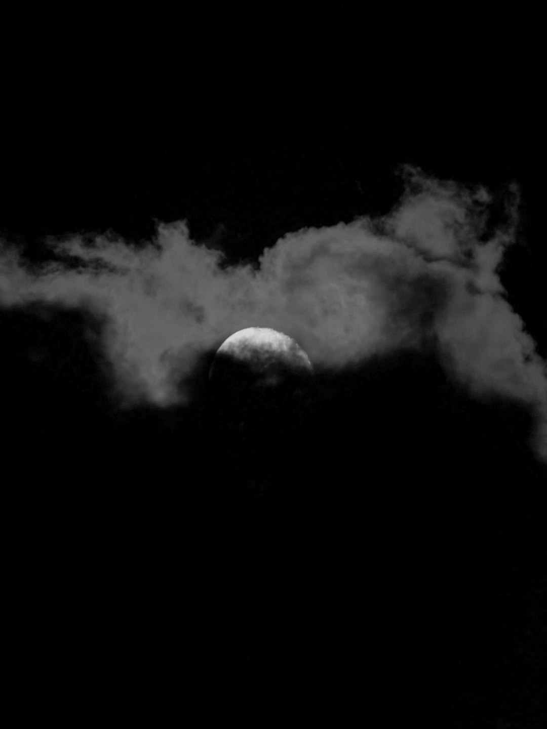 A black and white photograph of the moon peeking through clouds, creating an eerie silhouette against a dark sky. The moon’s glow is visible in shades of silver or gray, adding to its mysterious appearance. The background is black. –ar 3:4