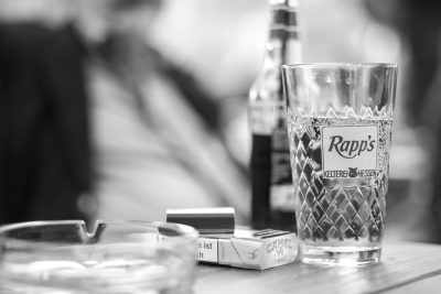 A black and white photo of "Raps's" glass with brandy, cigarette pack on the table in front of it, blurry background of people smoking cigarettes, cigarette box, cigarette holder, closeup shot, f/2 lens, Nikon D850 camera, shallow depth of field, natural light, retro style. --ar 128:85