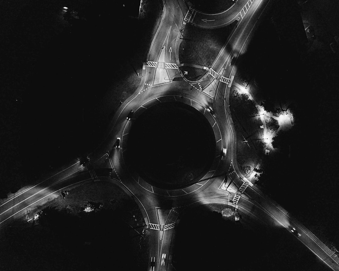 Black and white aerial view of an illuminated roundabout at night, showcasing the contrast between lighted areas on roads within the circle and darkness outside it. The scene captures the unique shape of a modern traffic intersection with streetlights creating dynamic patterns in black and white. –ar 64:51