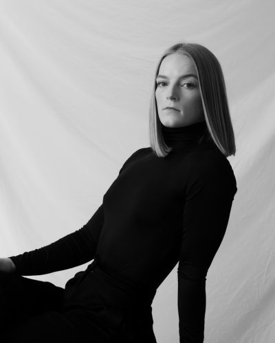 a black and white portrait of an adult female with straight blonde hair wearing a long sleeve turtle neck body suit, sitting on the ground looking at camera, posing for photoshoot, against a white backdrop, --ar 51:64