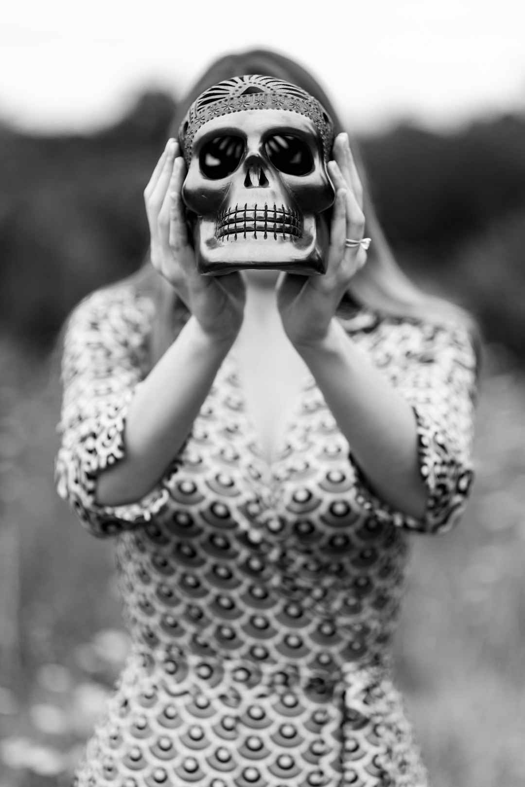 Black and white photography of an woman holding up her face mask with skull design, wearing patterned dress in nature. The subject is standing facing the camera directly –ar 85:128