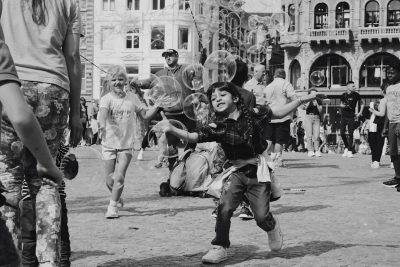 Black and white street photography of children playing with soap bubbles in the square. A crowded place, kids dressed up for a carnival party running around laughing, wearing costumes made from plastic bags in old buildings. A wide angle, high contrast style in the tradition of fujifilm superia street photography. --ar 128:85