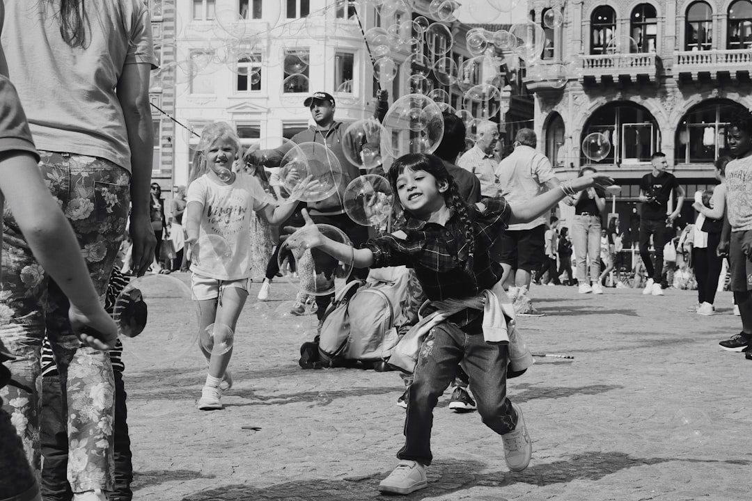 Black and white street photography of children playing with soap bubbles in the square. A crowded place, kids dressed up for a carnival party running around laughing, wearing costumes made from plastic bags in old buildings. A wide angle, high contrast style in the tradition of fujifilm superia street photography. –ar 128:85